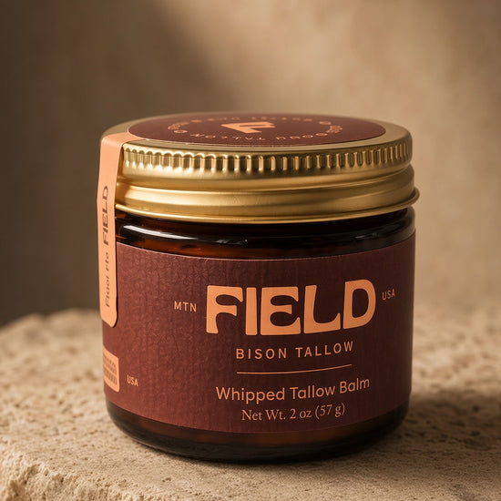 A jar of FIELD Bison Tallow Whipped Tallow Balm with a gold lid and dark brown label, resting on a textured stone surface with a warm, sandy background.