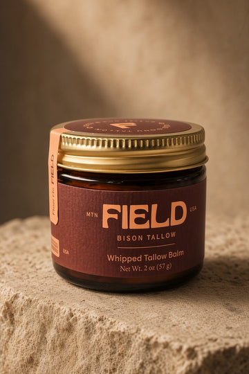 A jar of FIELD Bison Tallow Whipped Tallow Balm with a gold lid and dark brown label, resting on a textured stone surface with a warm, sandy background.
